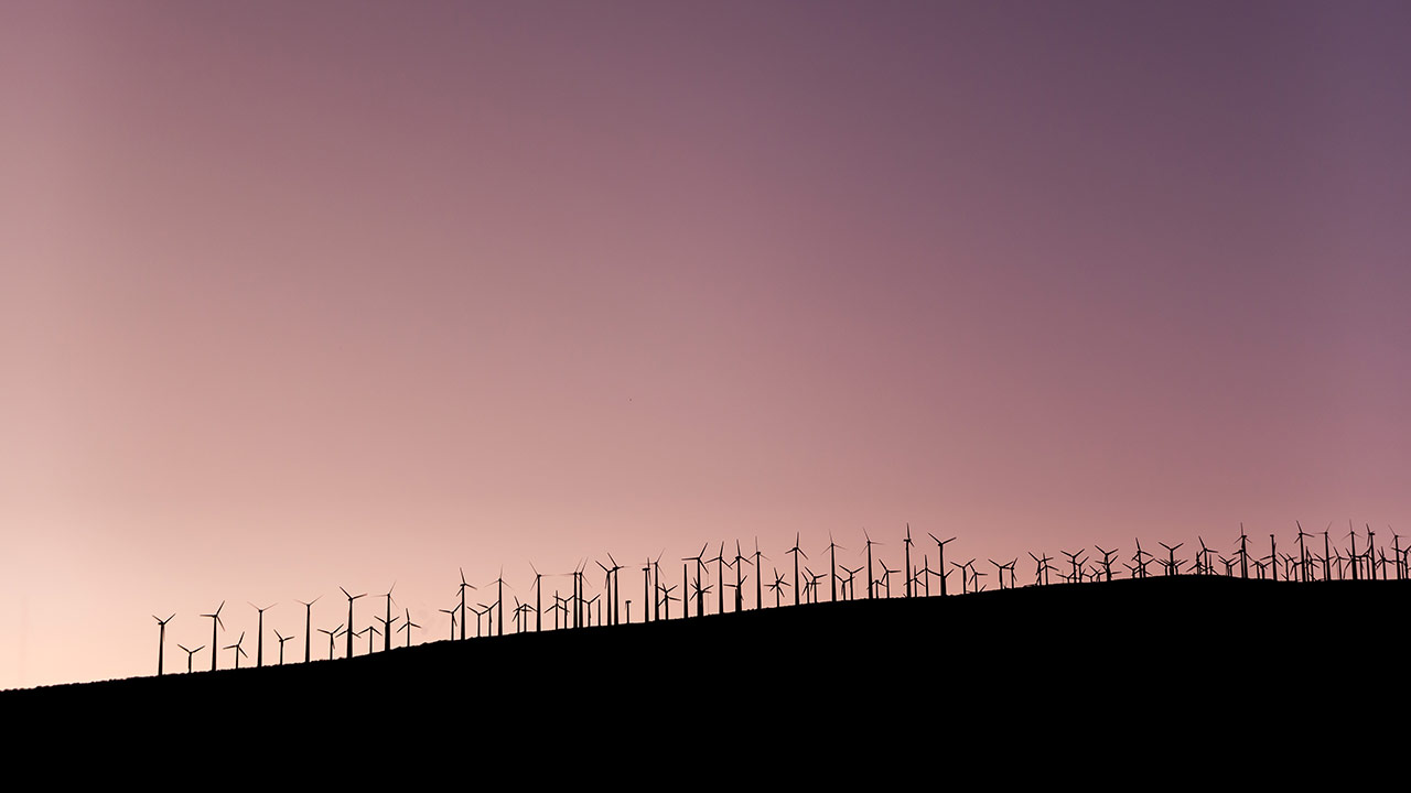 Das Foto zeigt zahlreiche Windkraftanlagen auf einem Hügel vor einem Sonnenuntergang.