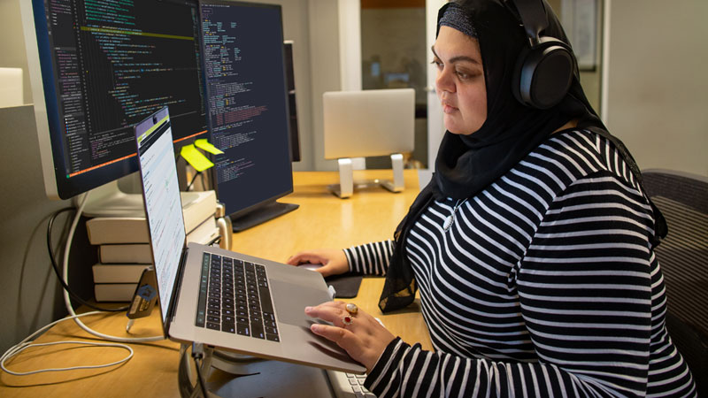 Eine Entwicklerin, die an ihrem Schreibtisch programmiert, Hijab und Kopfhörer trägt, hat ihren Arbeitsplatz mit einer Einrichtung mit mehreren Monitoren angepasst.